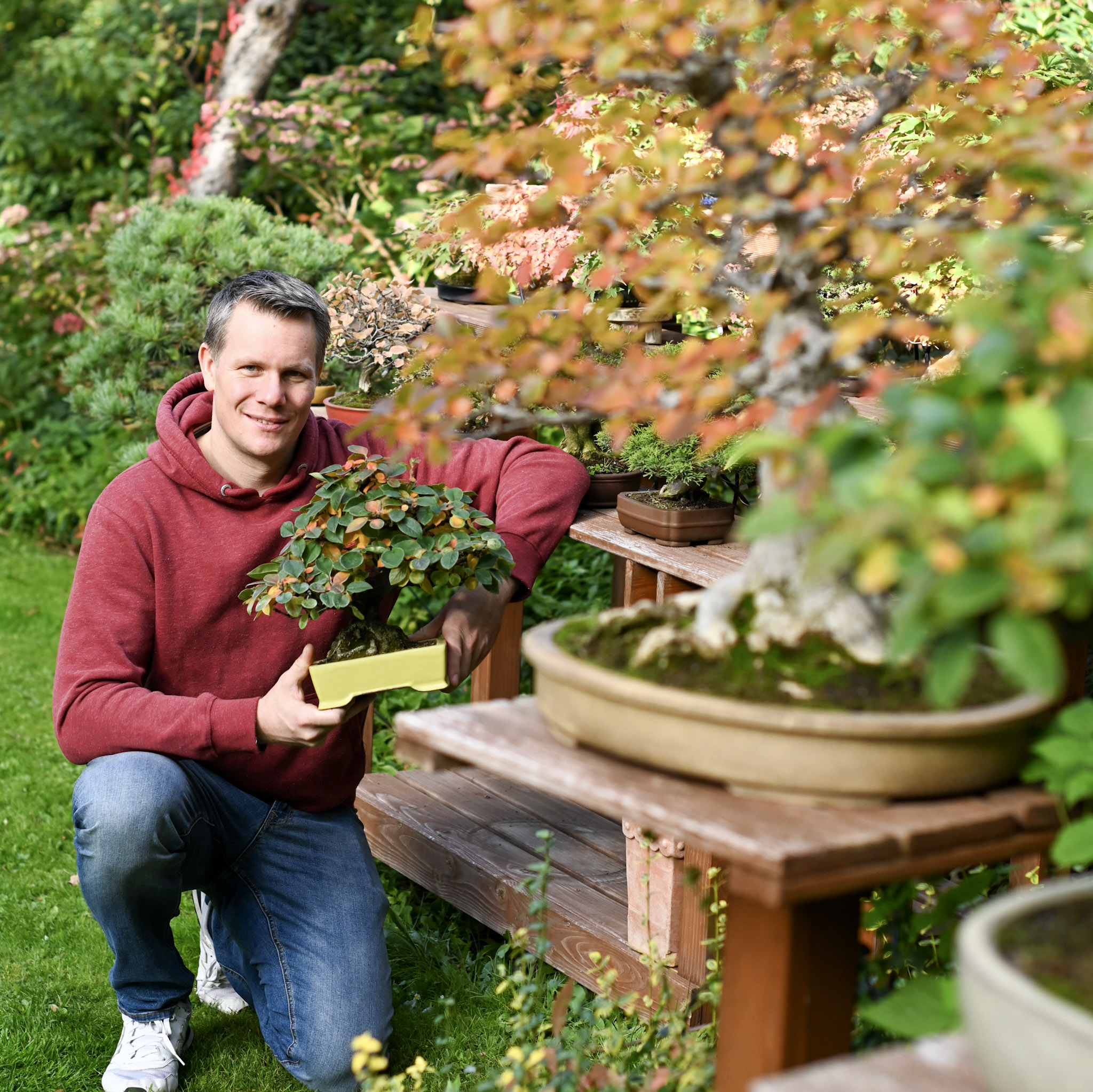 Der Inhaber von Ikigai Bonsai kniet mit einem Bonsai in der Hand vor einer schönen Bonsaikulisse